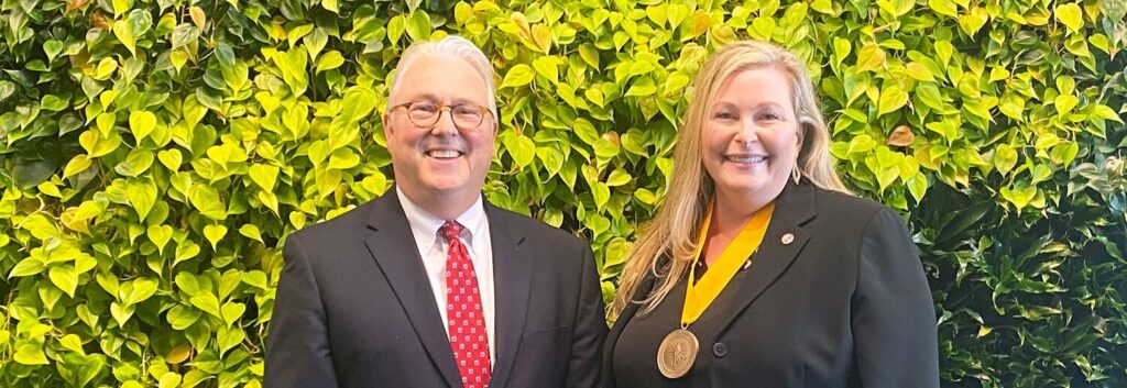 A woman in a dark suit has a golden award around her neck, with a yellow ribbon. A man in a suit stands next to her. They are smiling. They are standing in front of a solid wall of leaves.