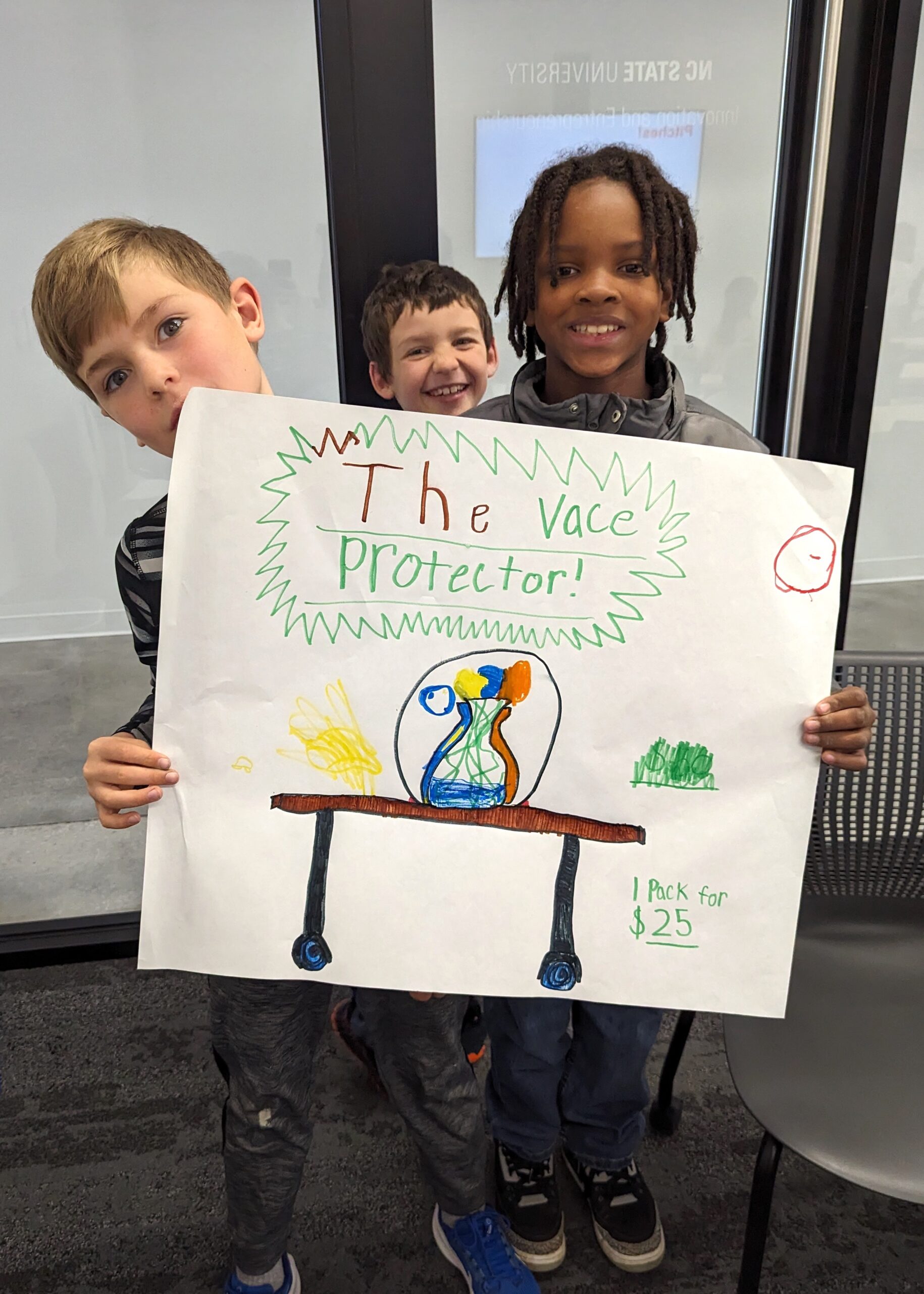 Three boys holding up a poster illustrating "the vace protector" invention, which is a globe that sits over a vase on a table.