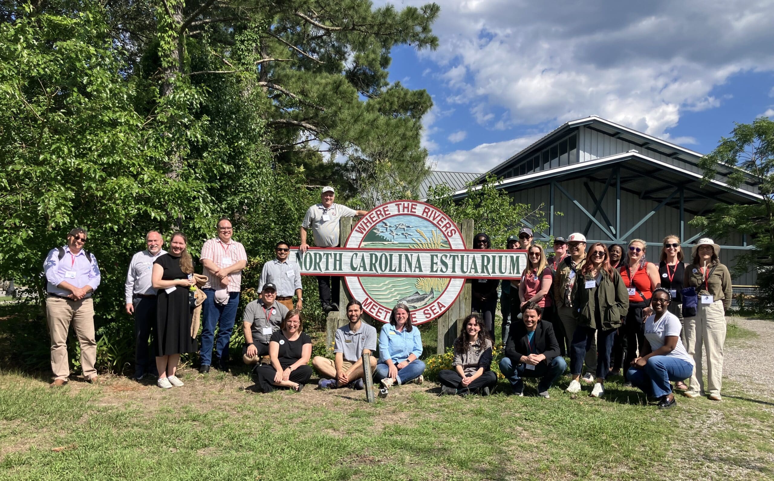 A group of adults pose next to a wooden sign for the North Carolina Estuarium, which has the motto Where the Rivers Meet the Sea. The group is outside next to the Estuarium's building